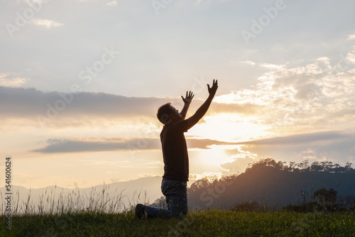 Young male kneeling down with hands open palm up praying to God on the mountain sunset background. photo