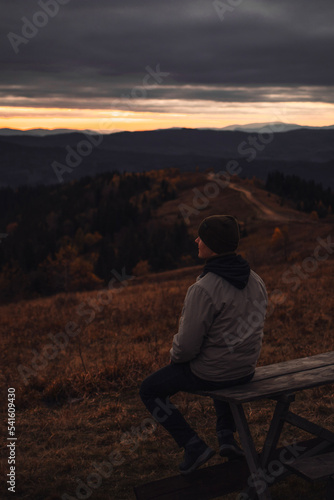 Young man is sitting at the bench and looking at sunset mountains view