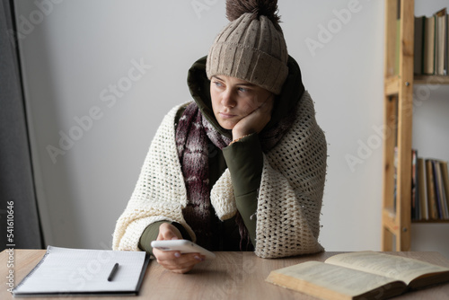 Fototapeta angry frozen woman warmly dressed in a cold house sitting at a table with mobile phone, problems with heating, energy crisis