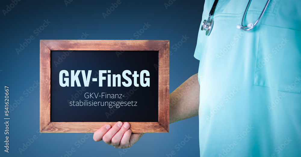 GKV-FinStG (GKV-Finanzstabilisierungsgesetz). Arzt zeigt Schild/Tafel mit Holz Rahmen. Hintergrund blau GKV-FinStG (GKV-Finanzstabilisierungsgesetz). Arzt zeigt Schild/Tafel mit Holz Rahmen. Hintergrund blau