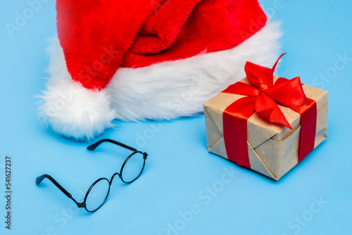 A Santa Claus hat, a Christmas gift tied with a red ribbon and round glasses on a blue background. A postcard for the new year. Copy space © Anastasiya Famina
