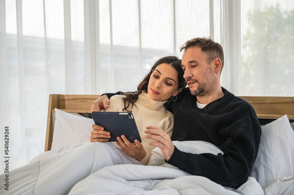 European couple man and woman smiling together looking at tablet mobile device sitting on bed under blanket. Male and female wearing warm winter clothing. European couple man and woman smiling together looking at tablet mobile device sitting on bed under blanket. Male and female wearing warm winter clothing.