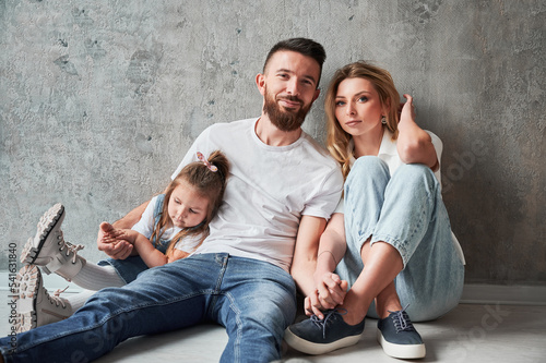 Loving parents and little child sitting by the wall in new apartment. Woman and man holding hands and looking at camera while cuddling with daughter in new home.