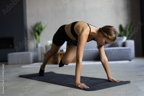 Enduring concentrated woman is doing working out at home and doing plank in front of her laptop, wearing sport outfit