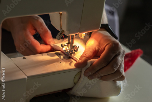Fototapeta Naklejka Na Ścianę i Meble -  Hands of experienced worker in the handmade industry, sewing red and white fabric on a sewing machine, the production of Santa red hat. Homemade sewing. Working tailor. Christmas hat. Close up.
