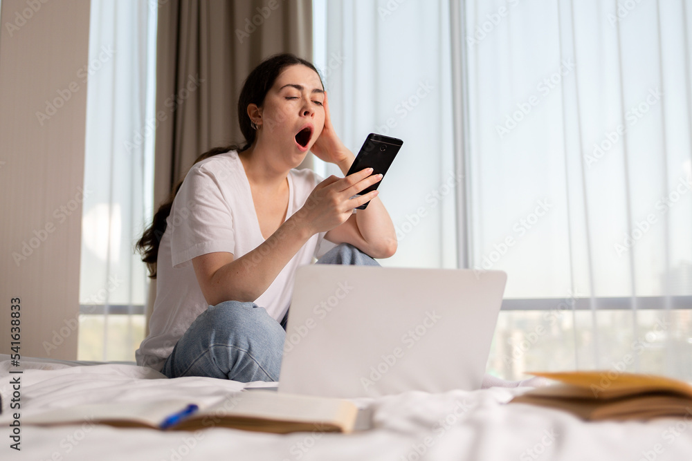 A young tired woman sits on the bed and yawns, using a smartphone. Next to it is a laptop and books. The concept of online education A young tired woman sits on the bed and yawns, using a smartphone. Next to it is a laptop and books. The concept of online education