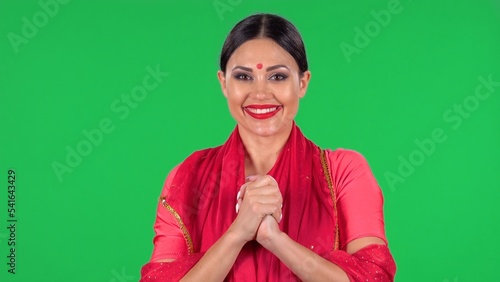 Portrait of a young Indian girl with red bindi dot in national classic red sari looking straight, smiles and folded palm to palm. A brunette with long hair and red lips on green screen at studio. photo