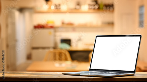 Laptop on wooden table against blurred kitchen with soft and warm light in background. Blank screen for your advertise design © Prathankarnpap