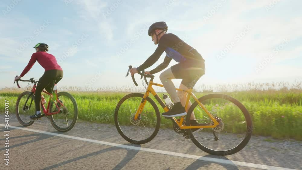 Side view of two professional cyclists riding on paved road during summertime. Happy friends in safety helmets enjoying endurance training on fresh air. Side view of two professional cyclists riding on paved road during summertime. Happy friends in safety helmets enjoying endurance training on fresh air.