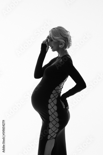 Beautiful short-haired pregnant woman in black dress with holes. Stylish girl in fashion studio shoot. Future mother expecting child. Black and white