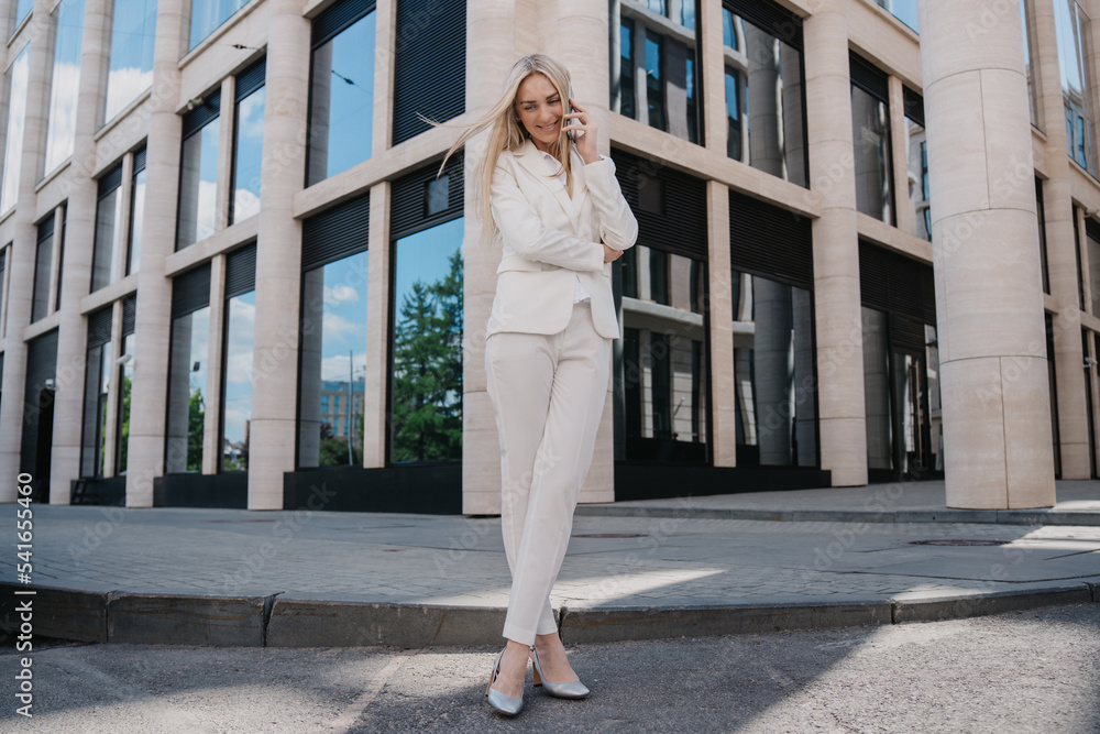 Pretty Italian businesswoman in white suit standing outside talking by phone, toothy smiling. Cheerful student received good news. Gorgeous Swedish female satisfied by her career. Business, education.