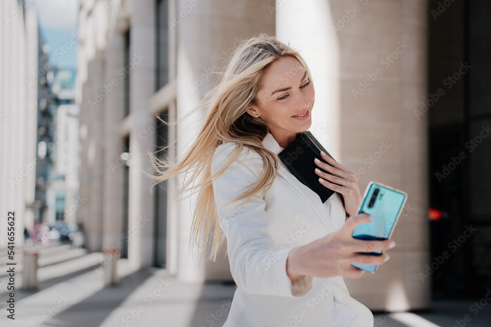 Gorgeous blonde woman in white suit walking by street smiling making selfie, talking by video call at summer day, going to work. Cheerful Italian successful businesswoman with flattering loose hair. Gorgeous blonde woman in white suit walking by street smiling making selfie, talking by video call at summer day, going to work. Cheerful Italian successful businesswoman with flattering loose hair.
