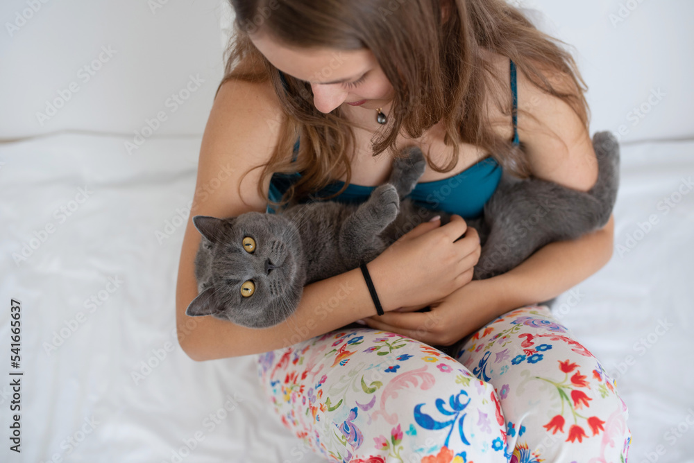 Young woman is holding her British shorthair cat like a baby while sitting on a bed Young woman is holding her British shorthair cat like a baby while sitting on a bed