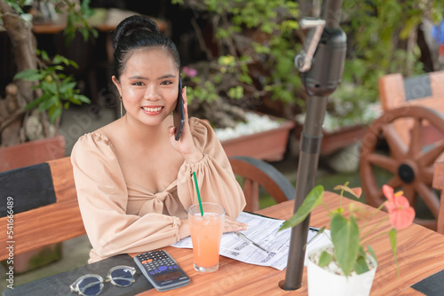 A smiling young woman sitting outside a fancy cafe is glad to hear the good news while talking on the phone with a client.