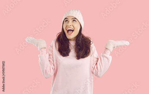 Happy excited woman in mittens rejoices, catches snowflakes and is impressed by winter. Cute Caucasian woman in warm mittens and hat joyfully spreads her hands and looks up isolated on pink background