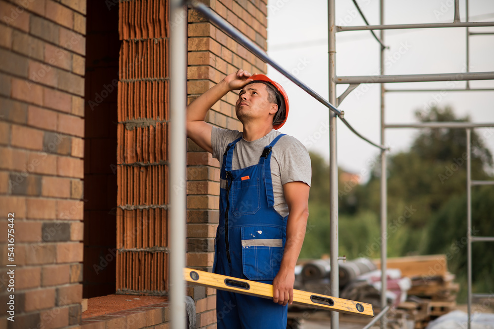 a male builder in blue overalls and a construction helmet stands near a window opening with a level, building a house