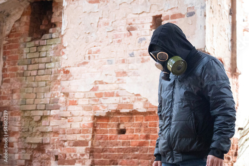 A guy in a gas mask stands against a brick wall and stares at the ground. A sad look in the photo