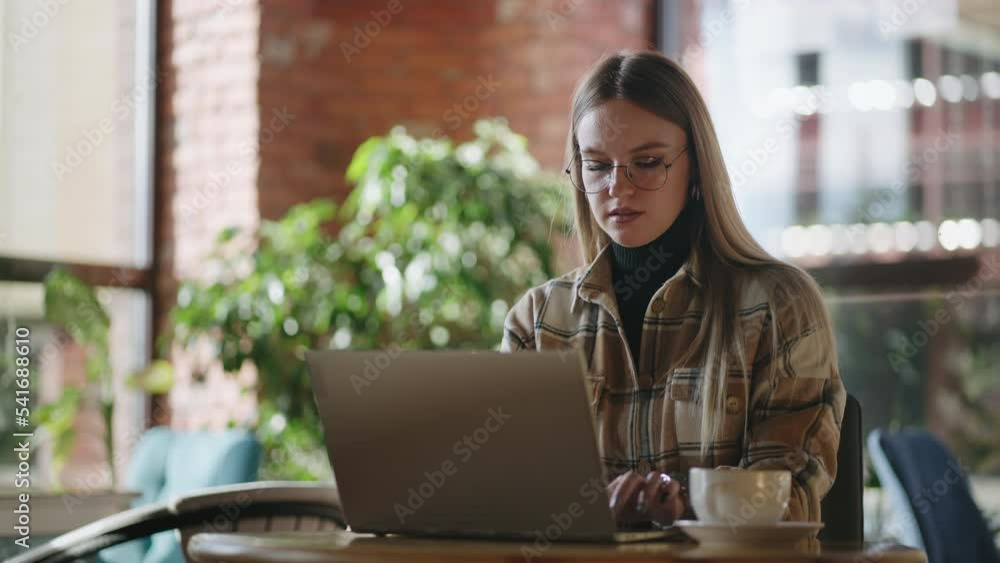 Serious blonde woman wearing eyeglasses is working on her laptop while sitting in a conference hall Serious blonde woman wearing eyeglasses is working on her laptop while sitting in a conference hall