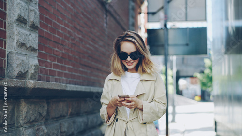 Pretty blond girl is using modern smartphone touching screen walking in city and smiling enjoying device. Young woman is wearing fashionable clothing and trendy sun glasses.