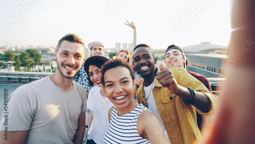 Cheerful African American girl is taking selfie with her friends, posing and laughing at rooftop party. Modern city is visible in background.