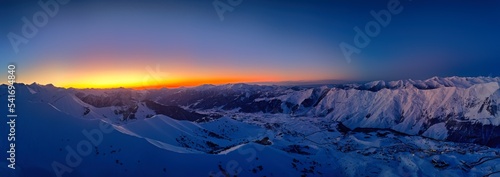 Wide aerial panorama of snowy mountain ridge on winter sunrise. Stunning mountains range covered with snow powder on ski resort at sunset. Caucasus mountain peaks skyline in the dusk golden hour.