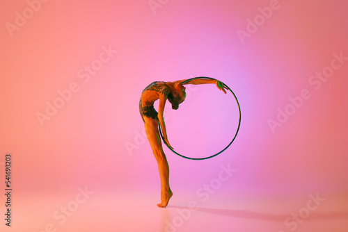 Young flexible teen girl rhythmic gymnast in motion, action isolated over pink background in neon light. Sport, beauty, competition, flexibility, active lifestyle