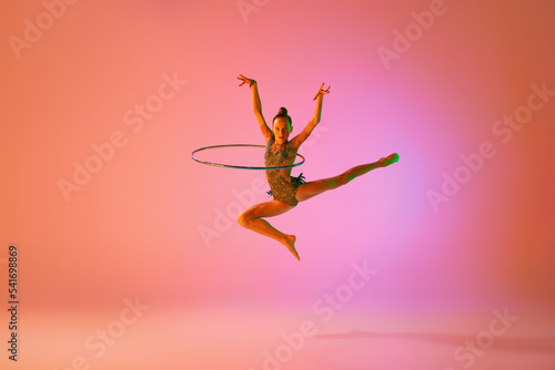 Young flexible teen girl rhythmic gymnast jumping isolated over pink background in neon light. Sport, beauty, competition, flexibility, active lifestyle