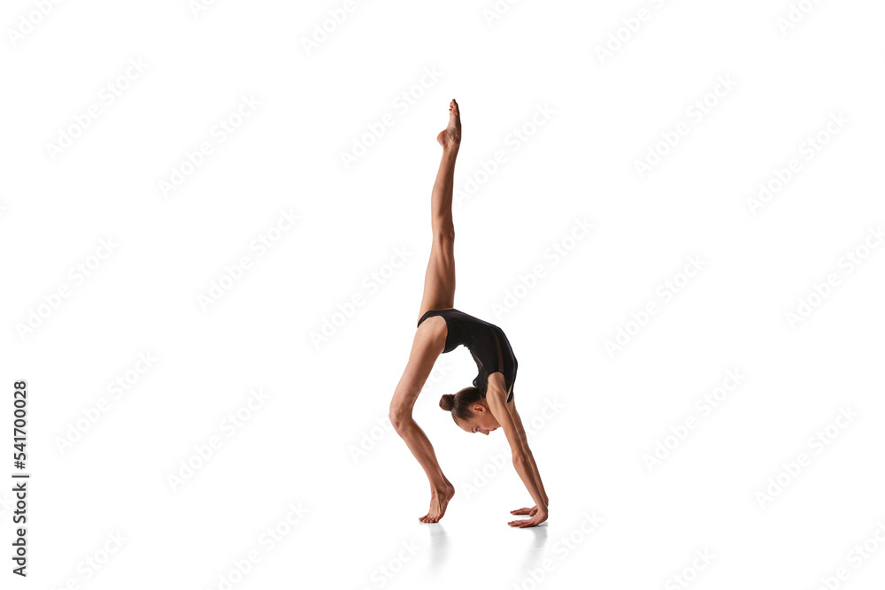 Balance. Portrait of junior gymnast in black sport swimsuit doing gymnastics excercises isolated over white background. Sport, skills, achievements