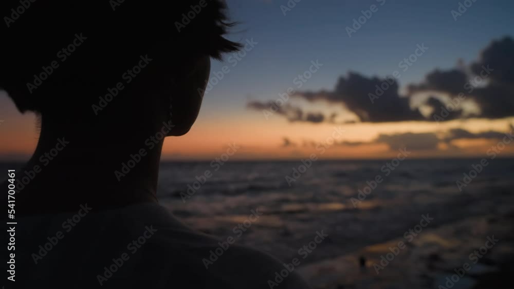 Silhouette from back of girl with short hair dreaming at seaside in evening light. Woman relaxing meditating alone on ocean beach at sunset sky background. Stress overcome. Film grain texture. Silhouette from back of girl with short hair dreaming at seaside in evening light. Woman relaxing meditating alone on ocean beach at sunset sky background. Stress overcome. Film grain texture.