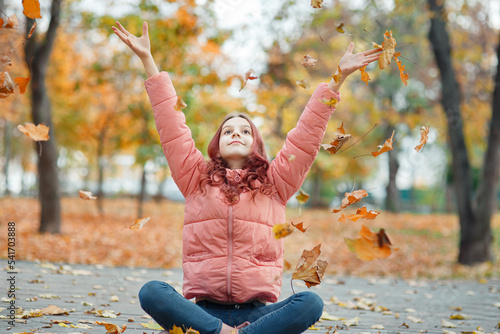 Young beautiful dreamy girl teenager sitting in autumn orange park in october  throwing maple leaves and smiling  hands up  seasonal outdoor lifestyle portrait  warm cozy mood 