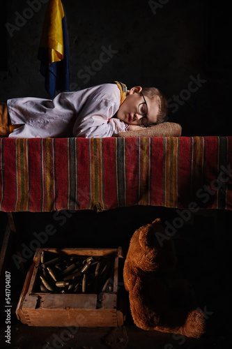 A child of war in Ukrainian clothes sleeps on a table covered with a bedspread. Under the table lies a box with cartridges and a teddy bear. Yellow and blue flag.