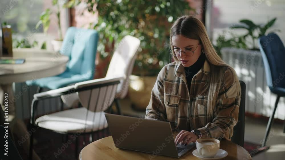 Serious blonde woman wearing eyeglasses is working on her laptop while sitting in a conference hall Serious blonde woman wearing eyeglasses is working on her laptop while sitting in a conference hall