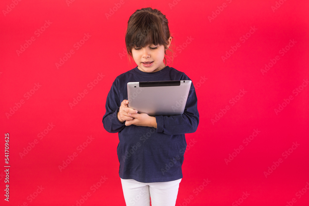 Smiling brunette-haired girl, wearing blue sweatshirt and white pants, holding and looking at digital tablet, on red background. Concept of call center, call, speak and listen.