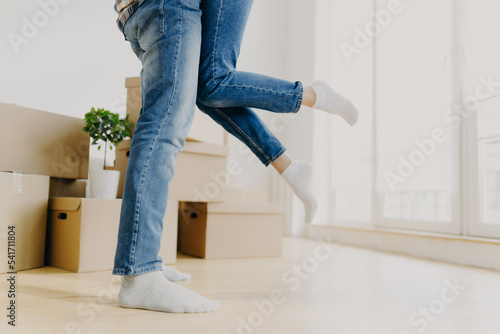 Happy unknown couple move in new abode, man lifts woman, wear jeans, pose in empty room with carton boxes around, start living seperate from parents rejoice buying apartment. Unrecognizable homeowners photo