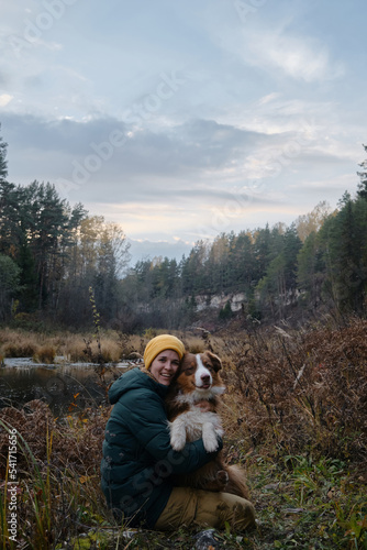 Spend time with animals in nature. Young Caucasian woman sitting happily with dog in autumn forest by river. Female pet owner hugs Australian Shepherd and smiles. Hiking with a dog.