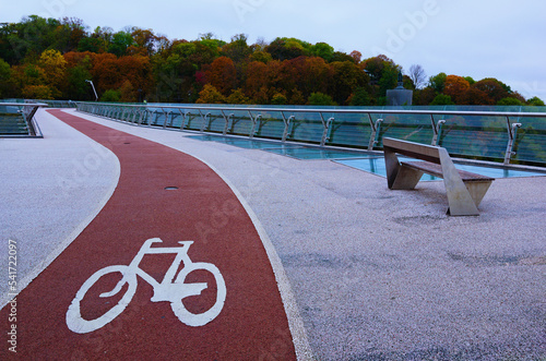 Scenic autumn landscape view of winding red color bike lane with white bicycle sign on the New Pedestrian Bridge in Kyiv, Ukraine. Beautiful autumn colored trees in the background