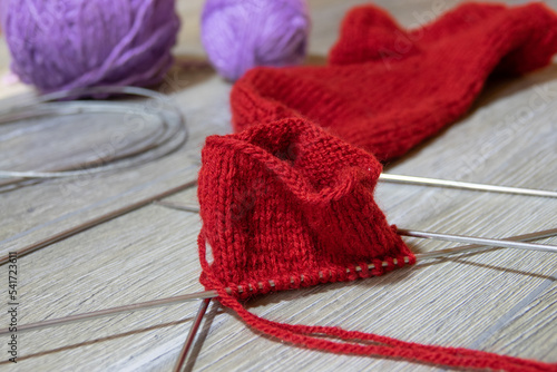 Sock in the process of knitting with needles and yarn of red color on a gray wooden table