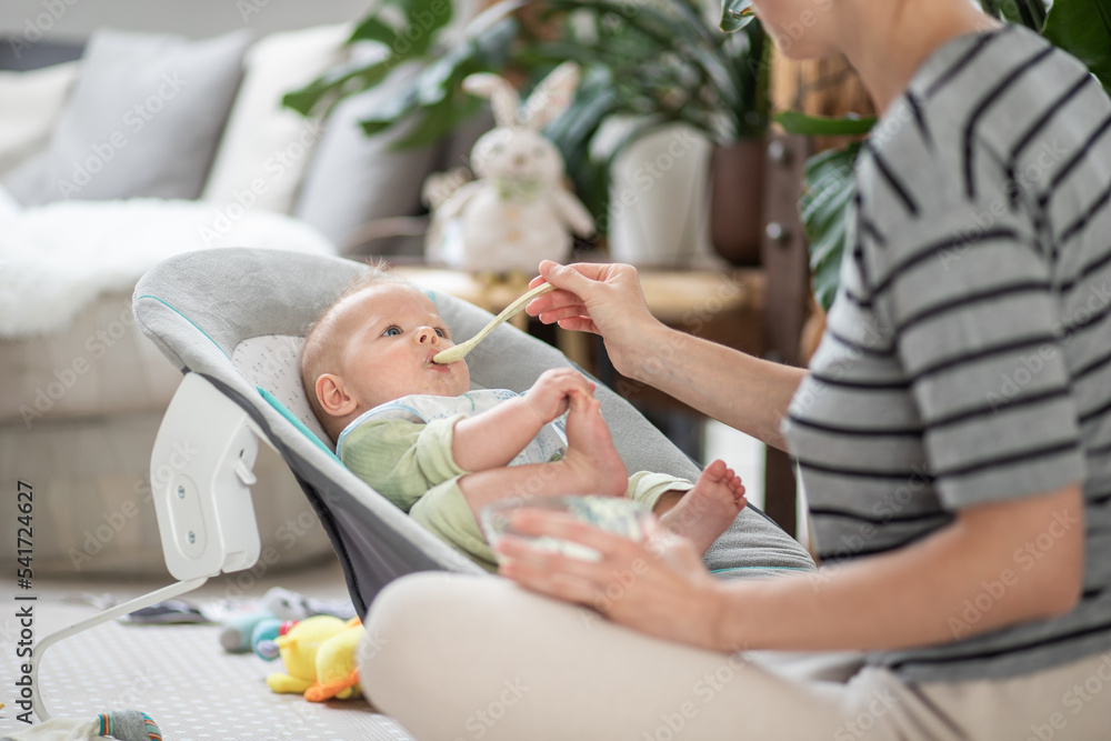 Mother spoon feeding her baby boy infant child in baby chair with fruit puree. Baby solid food introduction concept. Mother spoon feeding her baby boy infant child in baby chair with fruit puree. Baby solid food introduction concept.