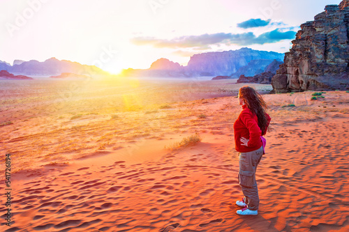 Rear view of a young female explorer in a red jacket staying on top of a dune enjoying a view of the picturesque red desert in Jordan