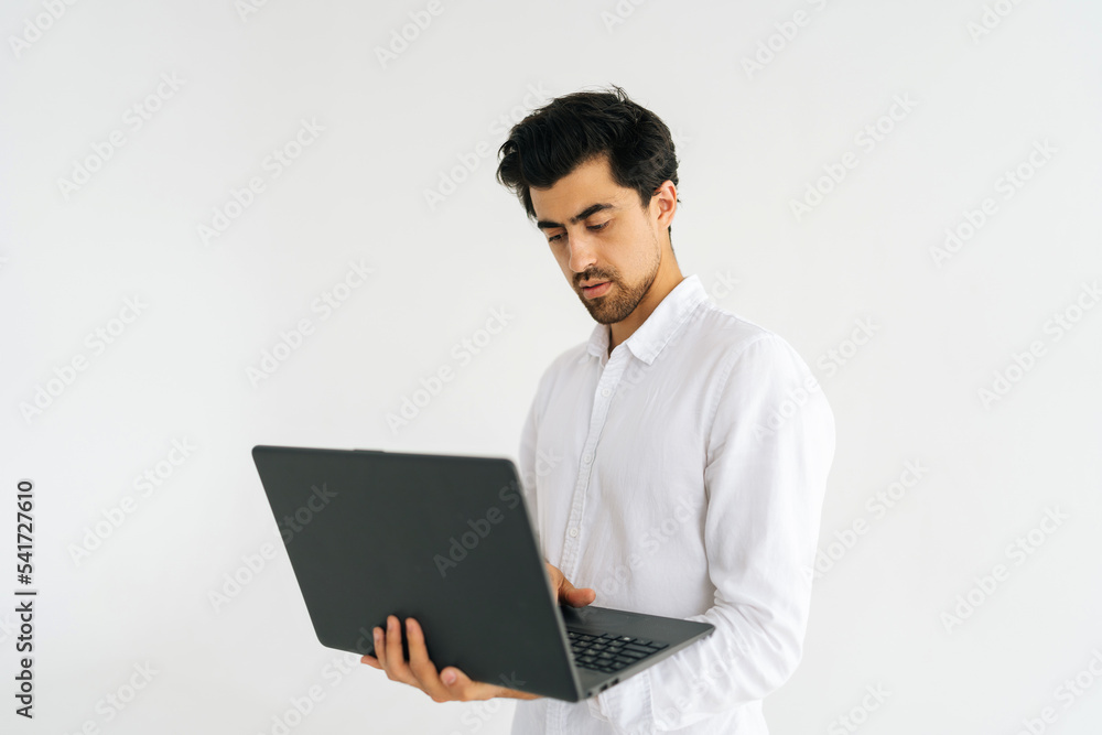 Studio shot of serious young man in shirt standing with laptop and typing on keyboard, doing freelance job, writing email on white isolated background. Focused male posing with pc looking at screen. Studio shot of serious young man in shirt standing with laptop and typing on keyboard, doing freelance job, writing email on white isolated background. Focused male posing with pc looking at screen.