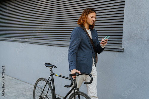 Ginger man using mobile phone while walking with bicycle on city street