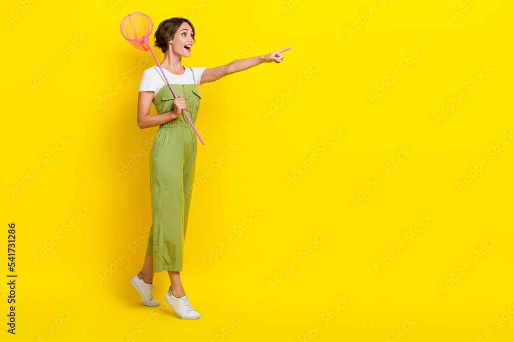 Full length size photo of active girl wear stylish khaki costume finger direct empty space hold butterfly net surprised isolated on yellow color background Full length size photo of active girl wear stylish khaki costume finger direct empty space hold butterfly net surprised isolated on yellow color background