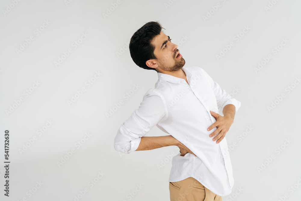 Studio portrait of unhealthy young man in shirt touching injured sore back, suffering lower lumbar discomfort, muscle pain of overwork, pinched nerve and grimacing on white isolated background. Studio portrait of unhealthy young man in shirt touching injured sore back, suffering lower lumbar discomfort, muscle pain of overwork, pinched nerve and grimacing on white isolated background.