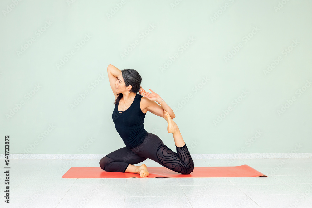 Fototapeta premium Sporty mature woman practicing yoga, sitting in Mermaid exercise, Eka Pada Rajakapotasana pose. Hispanic lady working out, wearing black sportswear, indoor. Physical mental health care concept.