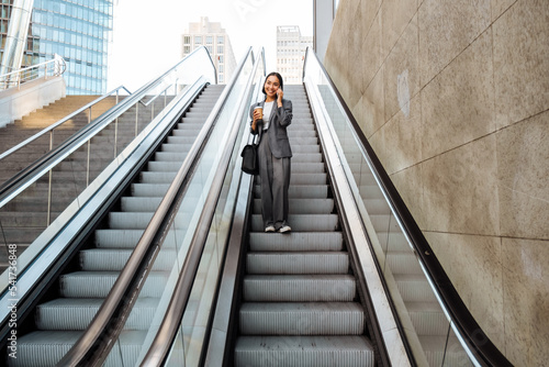 Young asian woman talking on cellphone and holding takeaway coffee while going down escalator outdoors