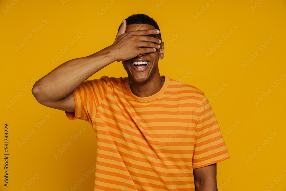 Afro man in striped orange shirt covering his eyes by hand standing isolated Afro man in striped orange shirt covering his eyes by hand standing isolated