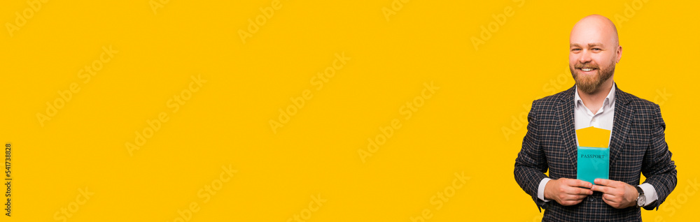 Banner size image of cheerful young bearded man in suit holding passport and flying tickets over yellow background. Banner size image of cheerful young bearded man in suit holding passport and flying tickets over yellow background.
