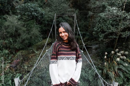 long haired caucasian young pretty woman in wool sweater on calm relaxed metal bridge crossing river near forest trees, mirror lake, new zealand photo