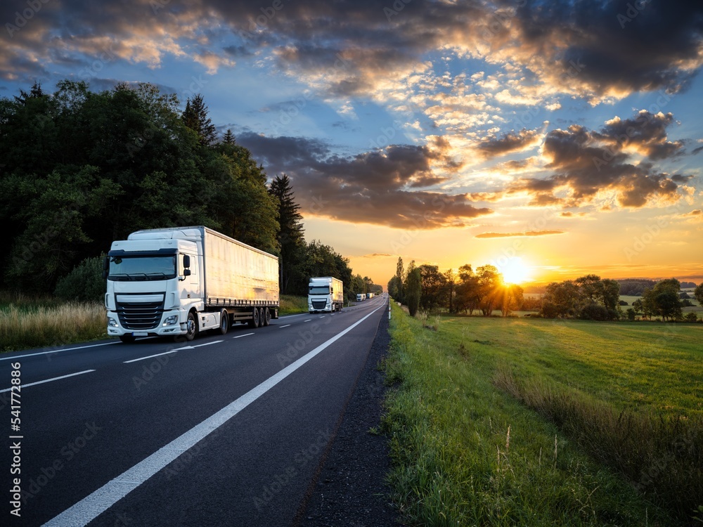 Two white trucks driving on the asphalt road in rural landscape at sunset