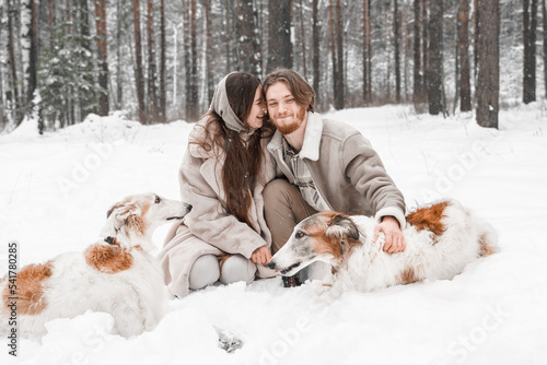 Fototapeta Naklejka Na Ścianę i Meble -  Love romantic young couple girl, guy in snowy cold winter forest walking with pet, dog of hunting breed russian borzoi. Sighthound, wolfhound owner. Having fun, laughing. Stylish fur coat, woolen hat